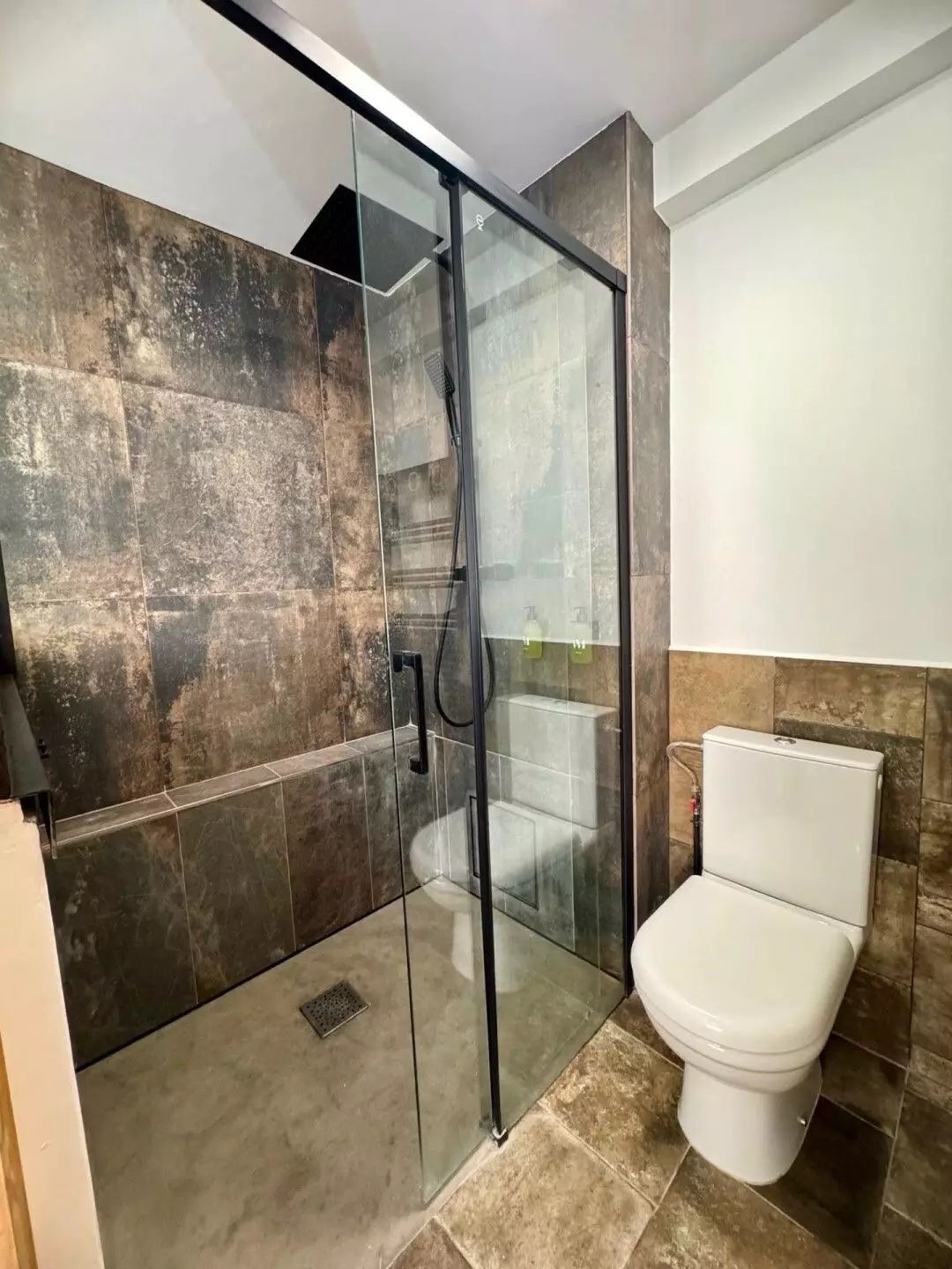 La grande douche avec paroi vitrée à cadre noir, pomme de pluie et douchette, carrelage sombre effet pierre, et les toilettes.